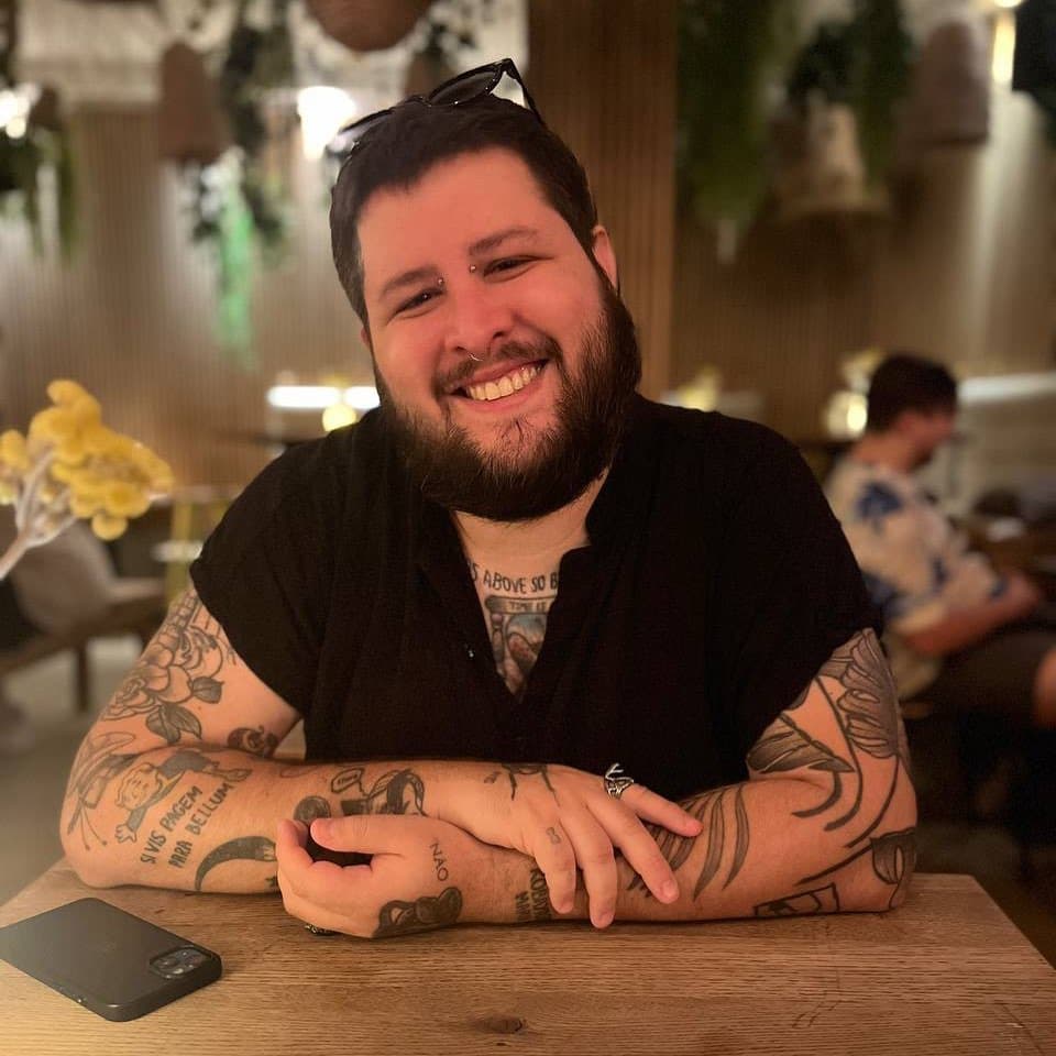 Foto de Angelo Dias sorrindo. Ele é um homem branco de cabelo e barba marrons escuro, com braços e peito tatuado, vestindo uma camisa preta.